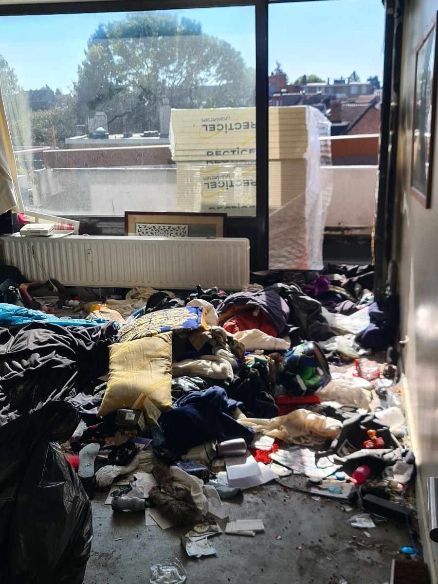 Séjour d’un appartement à Liège recouvert de vêtements, coussins, sacs et déchets jusqu’à la baie vitrée, avant le débarras